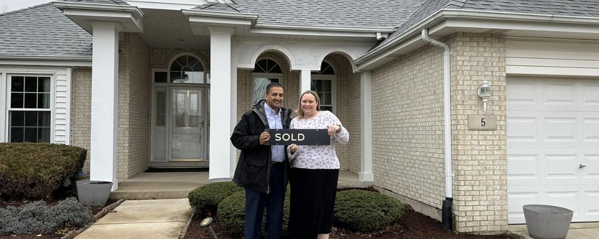 A couple standing in front of the house they bought with a "sold" sign